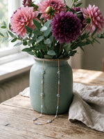 Lundholm Dahlia lange ketting met lichtblauwe kralen en bloemetjes goudkleurig over een groene vaas met dahlia's