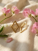 Lundholm Elvine geometrische oorbellen (hier goudkleurig) op crème linnen met roze lathyrus