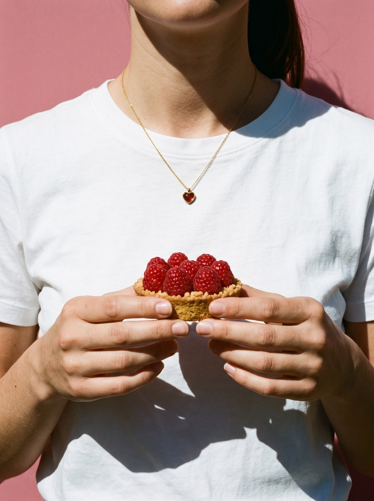 Vrouw in wit T-shirt draagt de Lundholm Emilia ketting met rood hartje goudkleurig, met een frambozentaartje