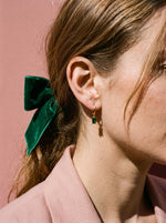 Vrouw met fluwelen strik draagt de Lundholm Gemma huggie oorringen met smaragdgroene zirkonia hanger goudkleurig