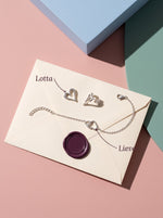 Lundholm Lotta open hartjes oorknopjes en Lieve armband met verbonden hartjes zilverkleurig op een envelop met bordeaux lakzegel