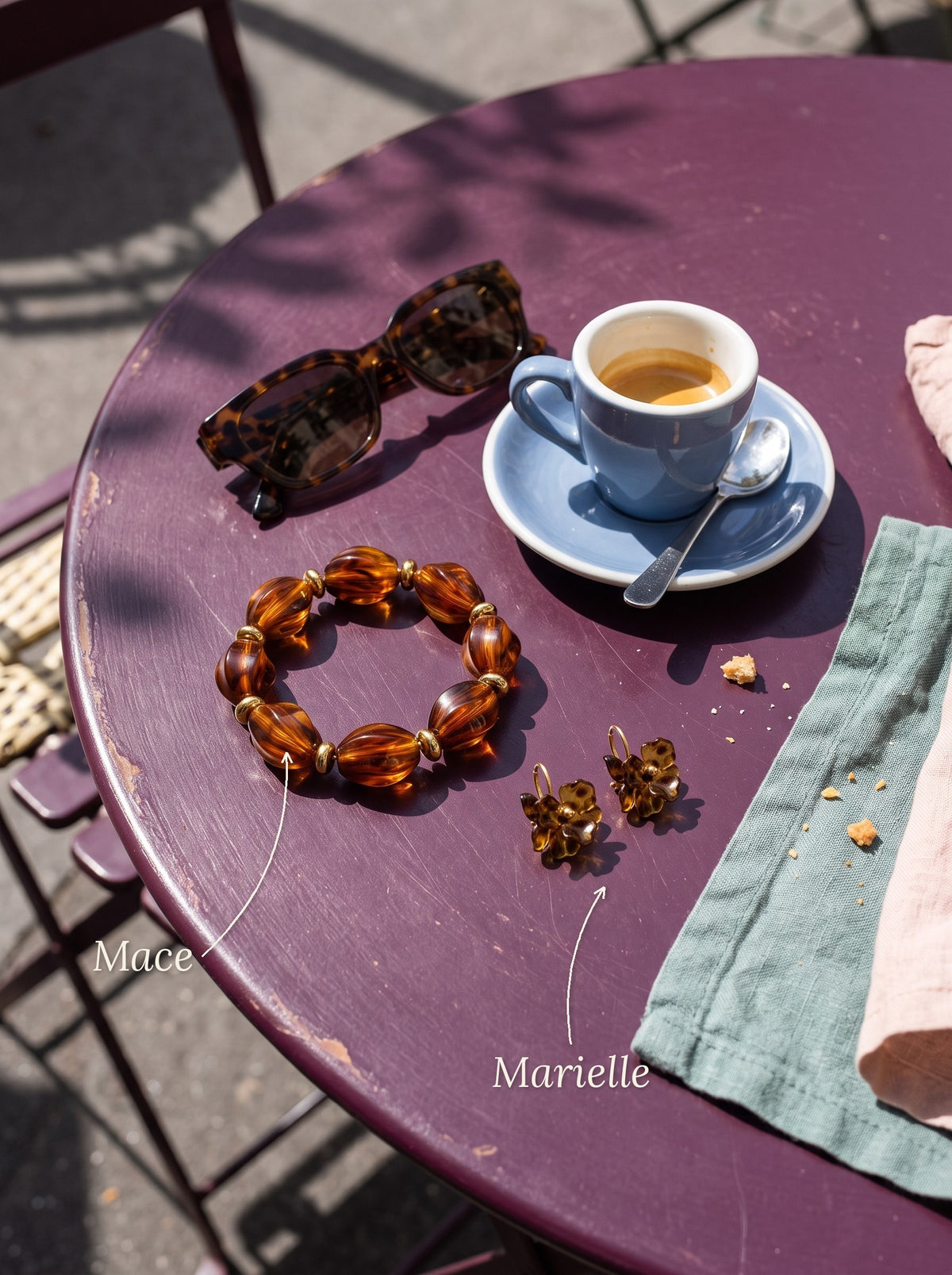 Lundholm Mace kralenarmband tortoise en Marielle bloem oorknopjes met amberkleurige steen, met hun namen erbij, op een cafétafel met espresso
