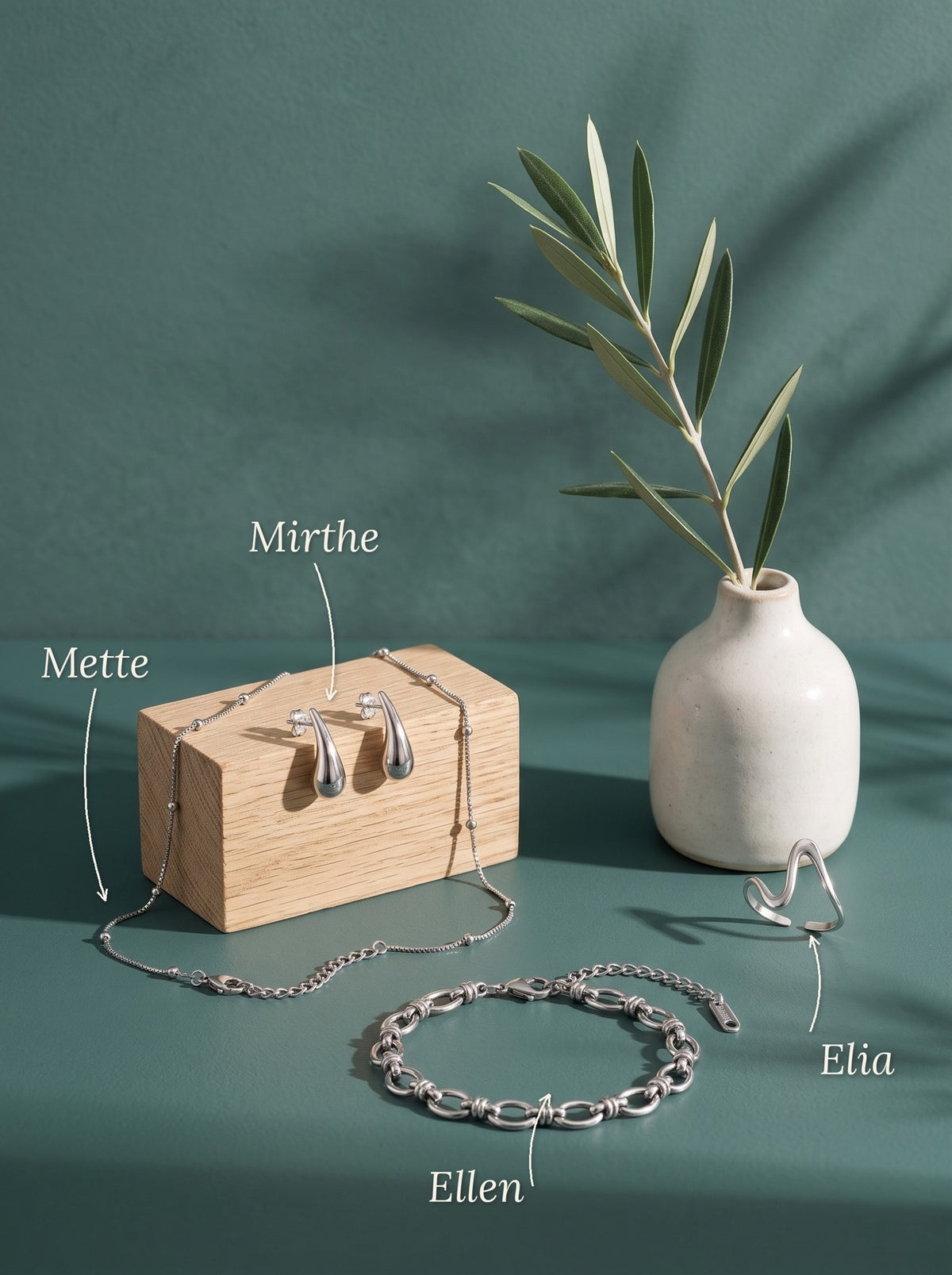 Lundholm Mette box chain ketting met bolletjes, Mirthe druppel oorbellen, Ellen schakelarmband en Elia golvende ring zilverkleurig bij een houten blokje en een wit vaasje met olijftak