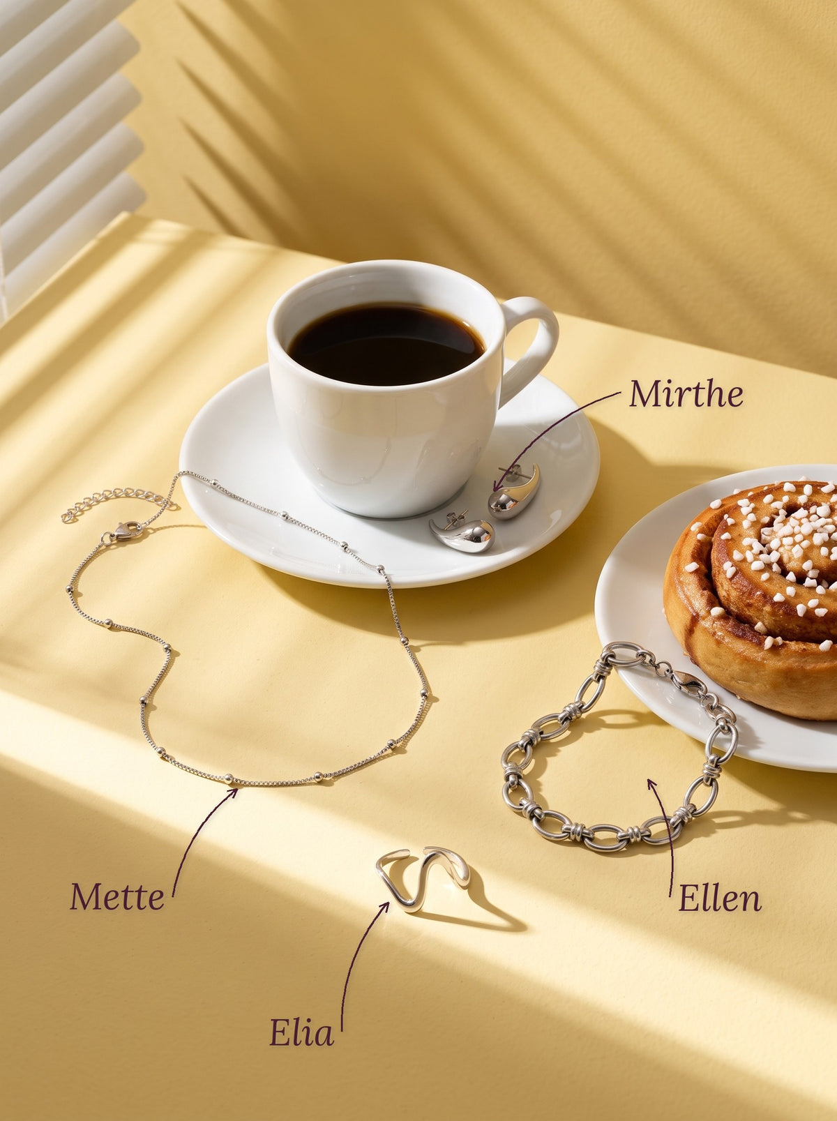 Lundholm Mette box chain ketting met bolletjes, Mirthe druppel oorbellen, Ellen schakelarmband en Elia golvende ring zilverkleurig naast een kop koffie en een kaneelbroodje