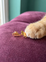 Kattenpootje reikt naar de Lundholm Minou kat oorknopjes goudkleurig op een pruimkleurig fluwelen kussen