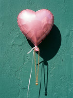 Lundholm Nanna hartjesketting goudkleurig met toggle sluiting aan een roze hartjesballon tegen een zeegroene muur