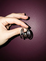 Hand met de Lundholm Nika triple hoop oorbellen zilverkleurig voor een pruimkleurige muur