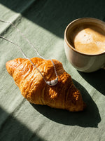 Lundholm Nilla ketting met croissant hanger zilverkleurig over een croissant naast een kop koffie