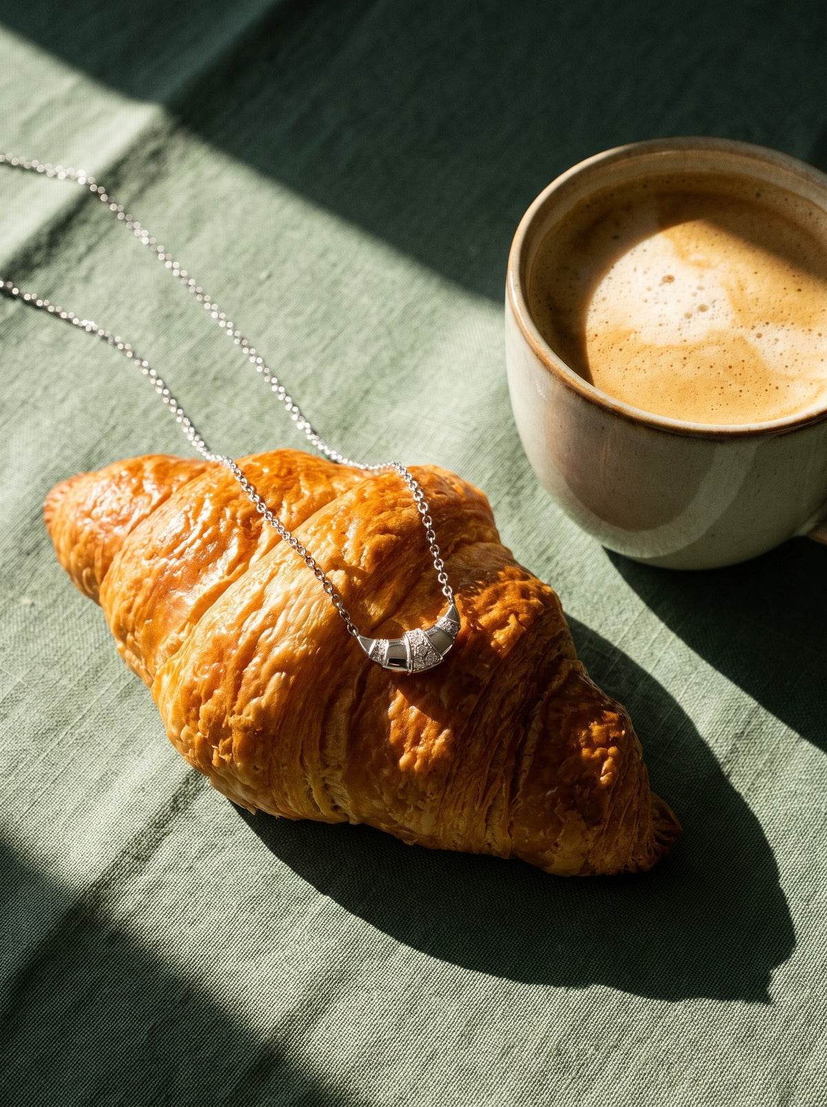 Lundholm Nilla ketting met croissant hanger zilverkleurig over een croissant naast een kop koffie