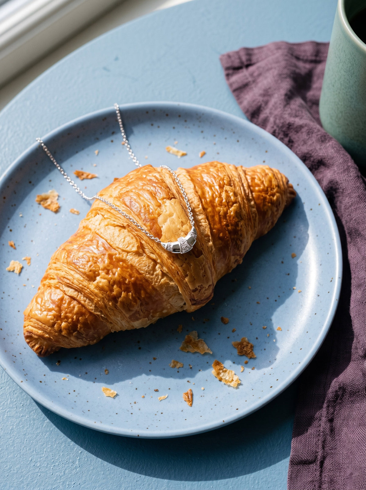 Lundholm Nilla ketting met croissant hanger zilverkleurig over een croissant op een blauw ontbijtbord