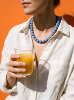 Vrouw in linnen blouse draagt de Lundholm Oslo kralenketting blauw met een glas sinaasappelsap