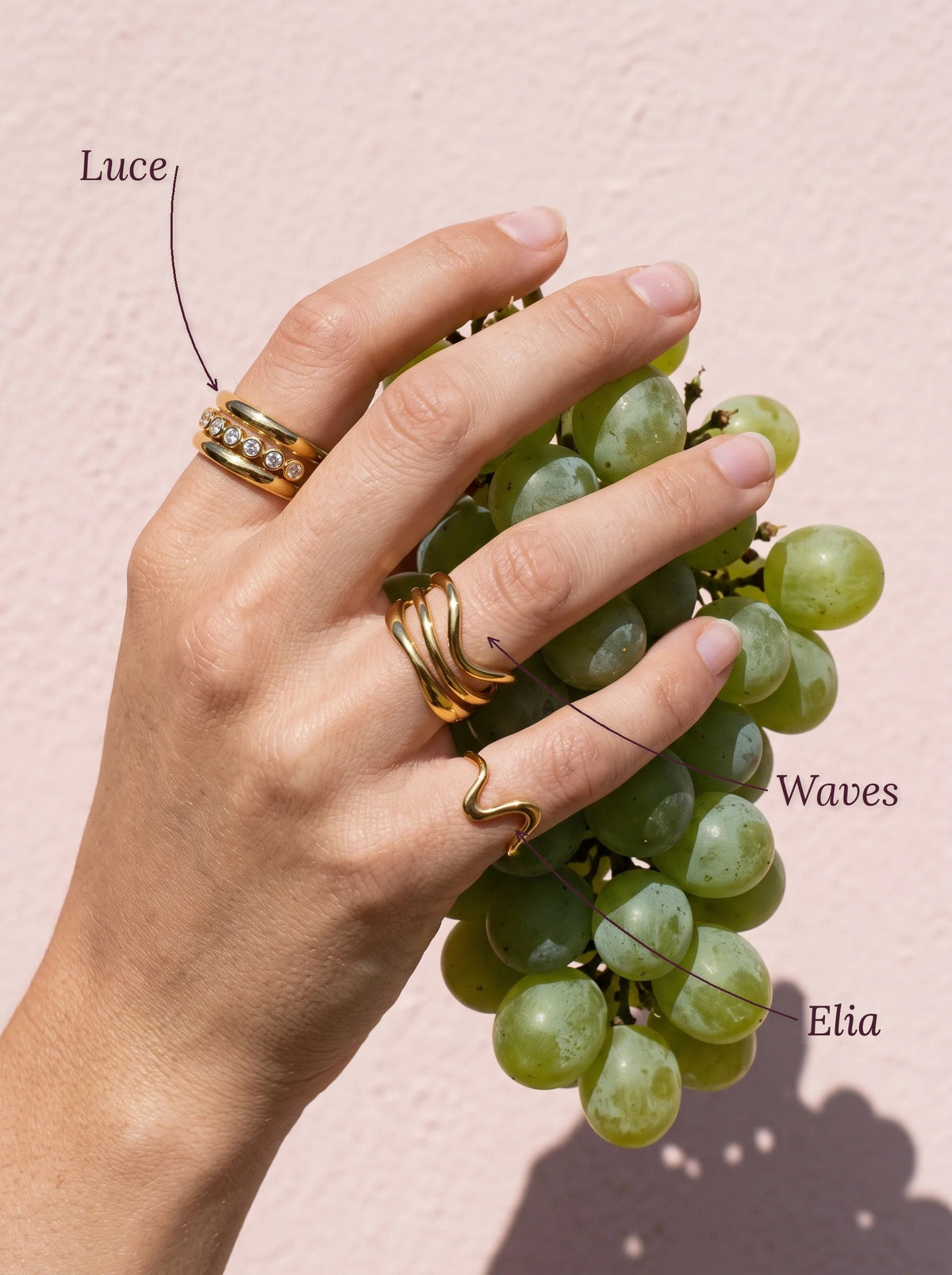 Hand met de Lundholm Luce ring met zirkonia, Waves golfring en Elia golvende ring goudkleurig en een tros witte druiven