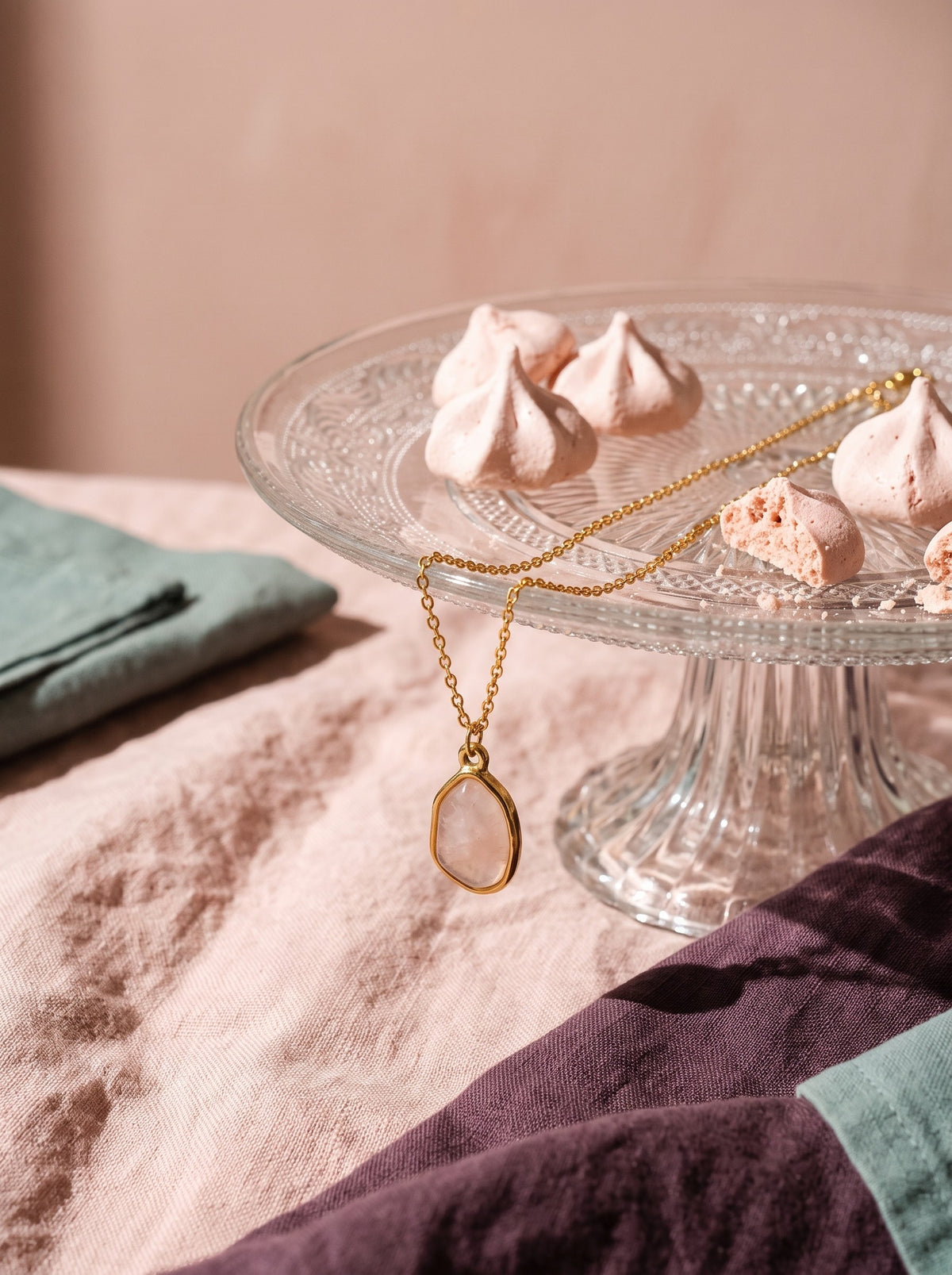 Lundholm Rosan ketting met rozenkwarts hanger goudkleurig over een glazen taartplateau met meringues