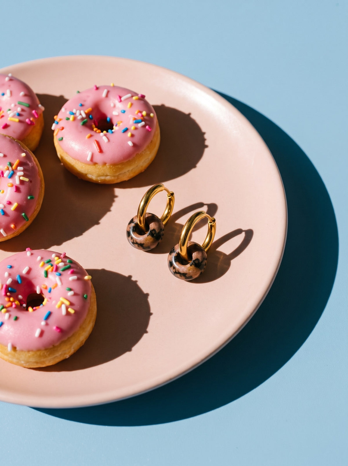 Lundholm Sally oorbellen met natuursteen donut goudkleurig op een bord met roze donuts