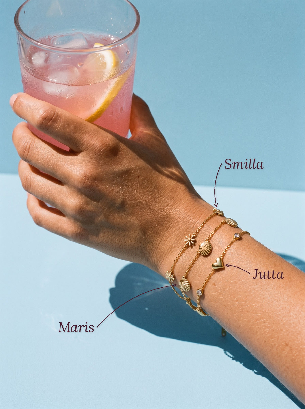 Lundholm Smilla madeliefjes armband, Maris schelpen armband en Jutta hartjes armband met zirkonia goudkleurig, met hun namen erbij, hand met roze limonade