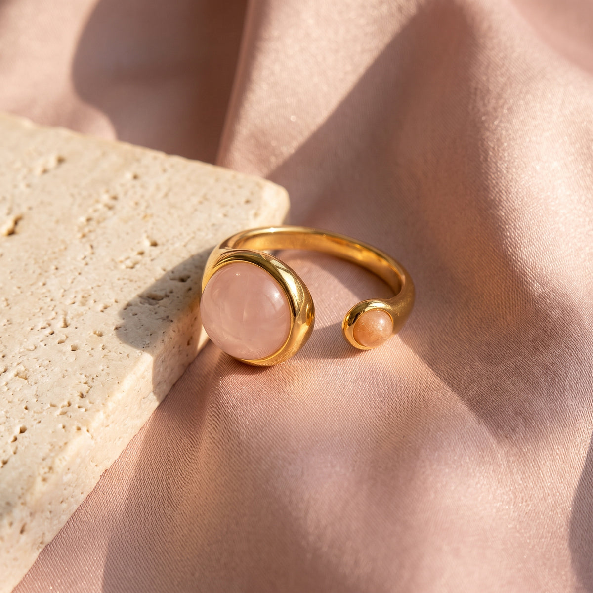 Lundholm Sophia ring met rozenkwarts en zonnesteen — flatlay op roze satijn en travertin