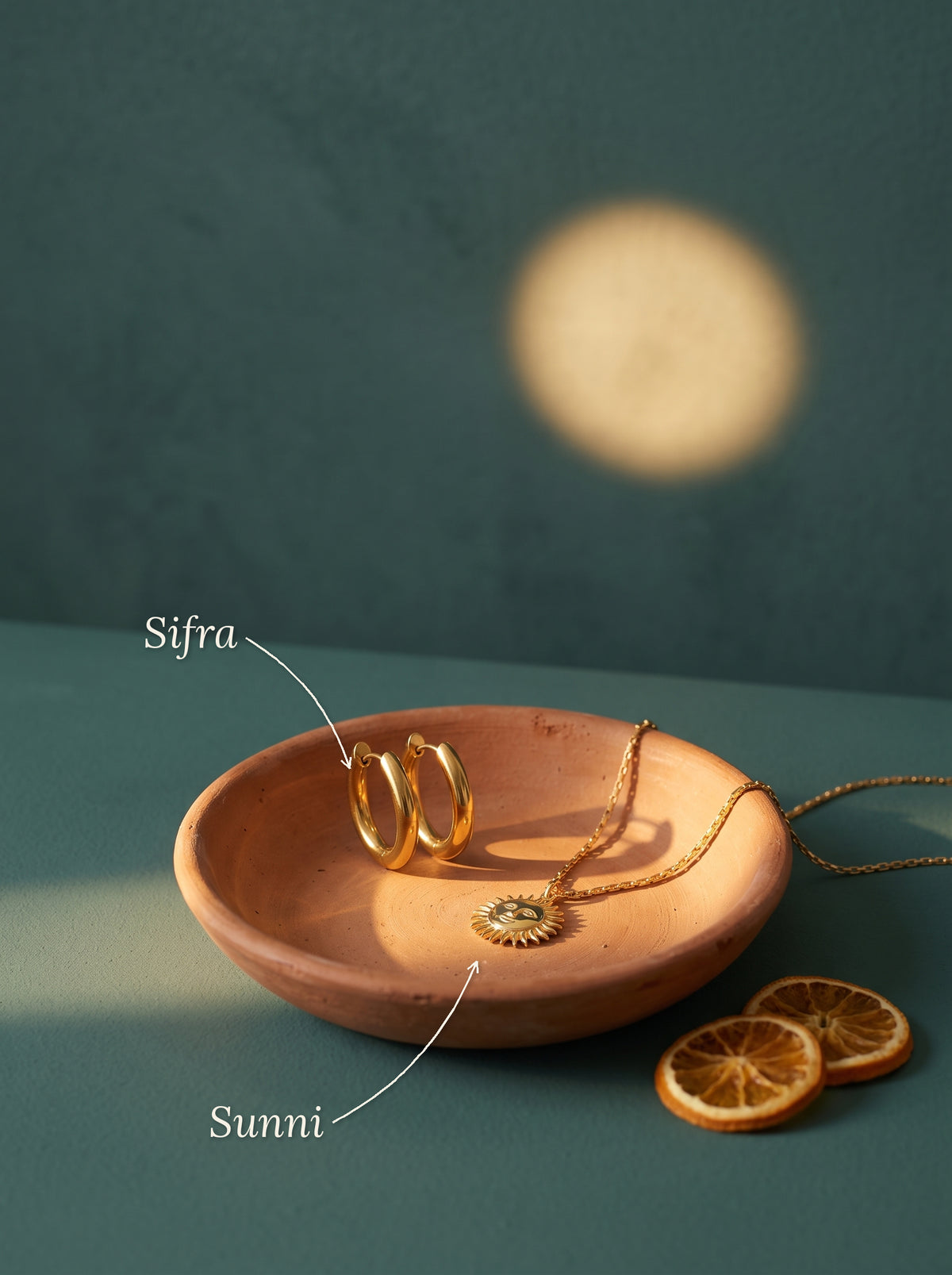 Lundholm Sunni ketting met zon hanger en Sifra ovale hoops goudkleurig in een terracotta schaal met gedroogde sinaasappel