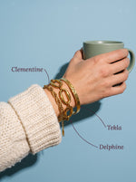 Lundholm Tekla gehamerde bangle, Clementine gedraaide schakelarmband en Delphine grove schakelarmband goudkleurig om de pols, met hun namen erbij