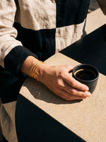 Lundholm Tindra open bangle met drie banen goudkleurig gedragen, hand om een espressokopje