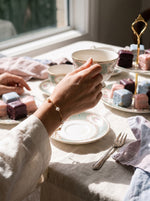 Hand met de Lundholm Vivienne parelarmband goudkleurig tilt een porseleinen theekopje op bij een high tea