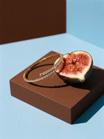 Lundholm Yael zirkonia tennisarmband bangle goudkleurig op een bruin doosje naast een halve vijg